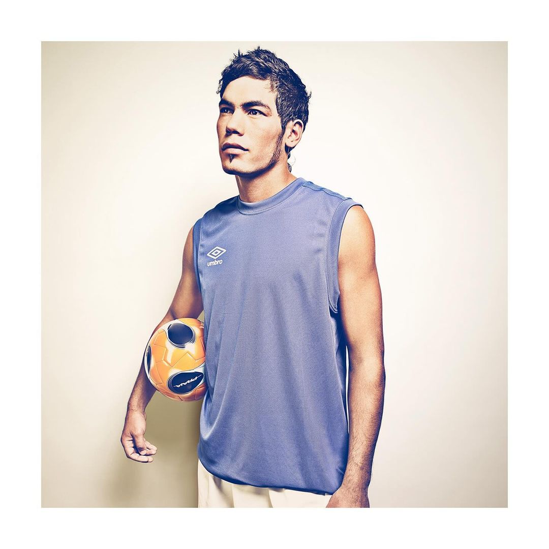 A male athlete in a blue tank top holds a soccer ball, looking upwards against a beige backdrop in a studio setting with soft lighting.