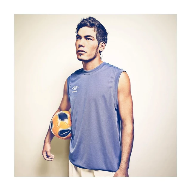 A male athlete in a blue tank top holds a soccer ball, looking upwards against a beige backdrop in a studio setting with soft lighting.