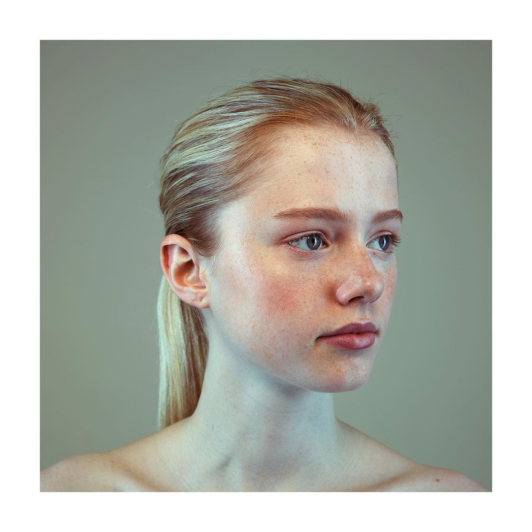Close-up portrait of a young woman with blonde hair pulled back in a ponytail, looking to the side against a neutral gradient background.