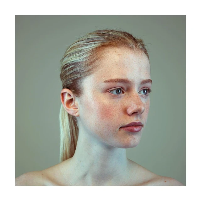 Close-up portrait of a young woman with blonde hair pulled back in a ponytail, looking to the side against a neutral gradient background.