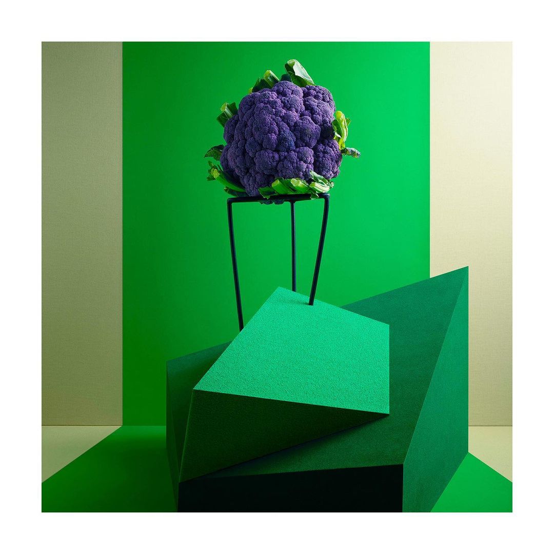 Vibrant purple cauliflower atop a black stand, geometric shapes below, against a green and beige backdrop. Still life food photography.