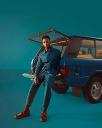 Man wearing a blue suit and brown shoes sitting on the open tailgate of a classic blue car against a matching teal studio backdrop.