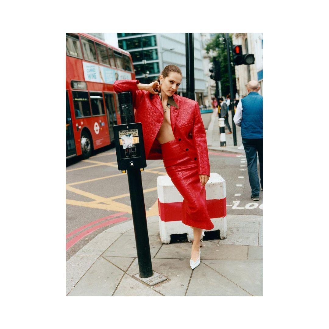 Adult female fashion model in a red leather jacket and skirt posing on a city street next to a crosswalk signal with a red double-decker bus.