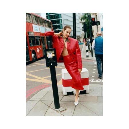 Adult female fashion model in a red leather jacket and skirt posing on a city street next to a crosswalk signal with a red double-decker bus.