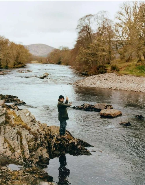 A man stands on rocks fishing in a river, wearing a dark coat and hat, surrounded by trees and a cloudy sky, in a natural setting.