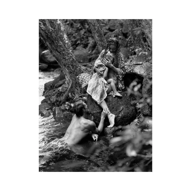 Monochrome image of a mother breastfeeds her baby on rocks by a stream, a man stands in the water nearby, creating a nature scene.