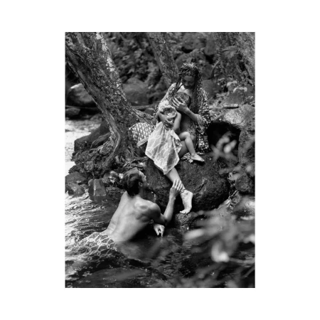 Monochrome image of a mother breastfeeds her baby on rocks by a stream, a man stands in the water nearby, creating a nature scene.