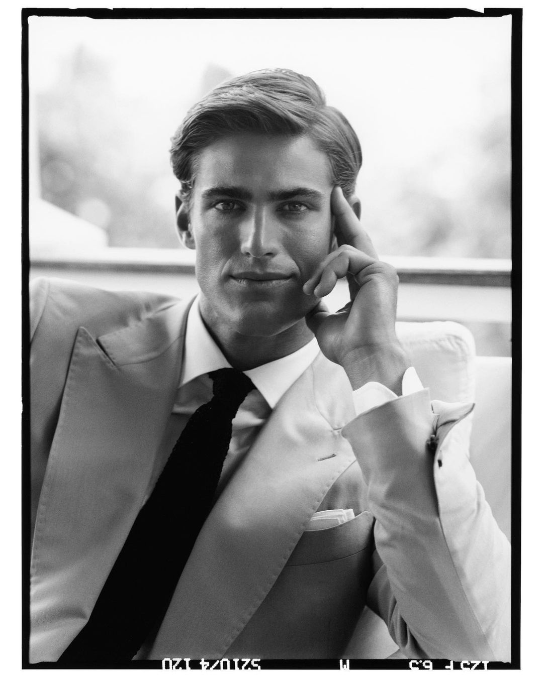 Portrait of a man in a suit and tie, hand to his head, sitting in an indoor environment, captured in black and white.