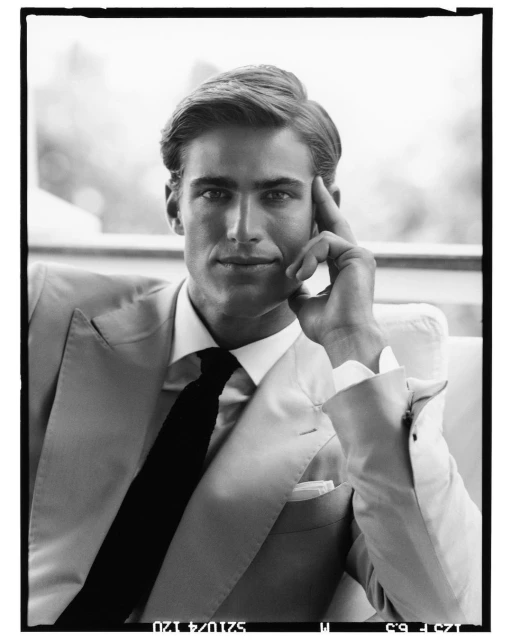 Portrait of a man in a suit and tie, hand to his head, sitting in an indoor environment, captured in black and white.