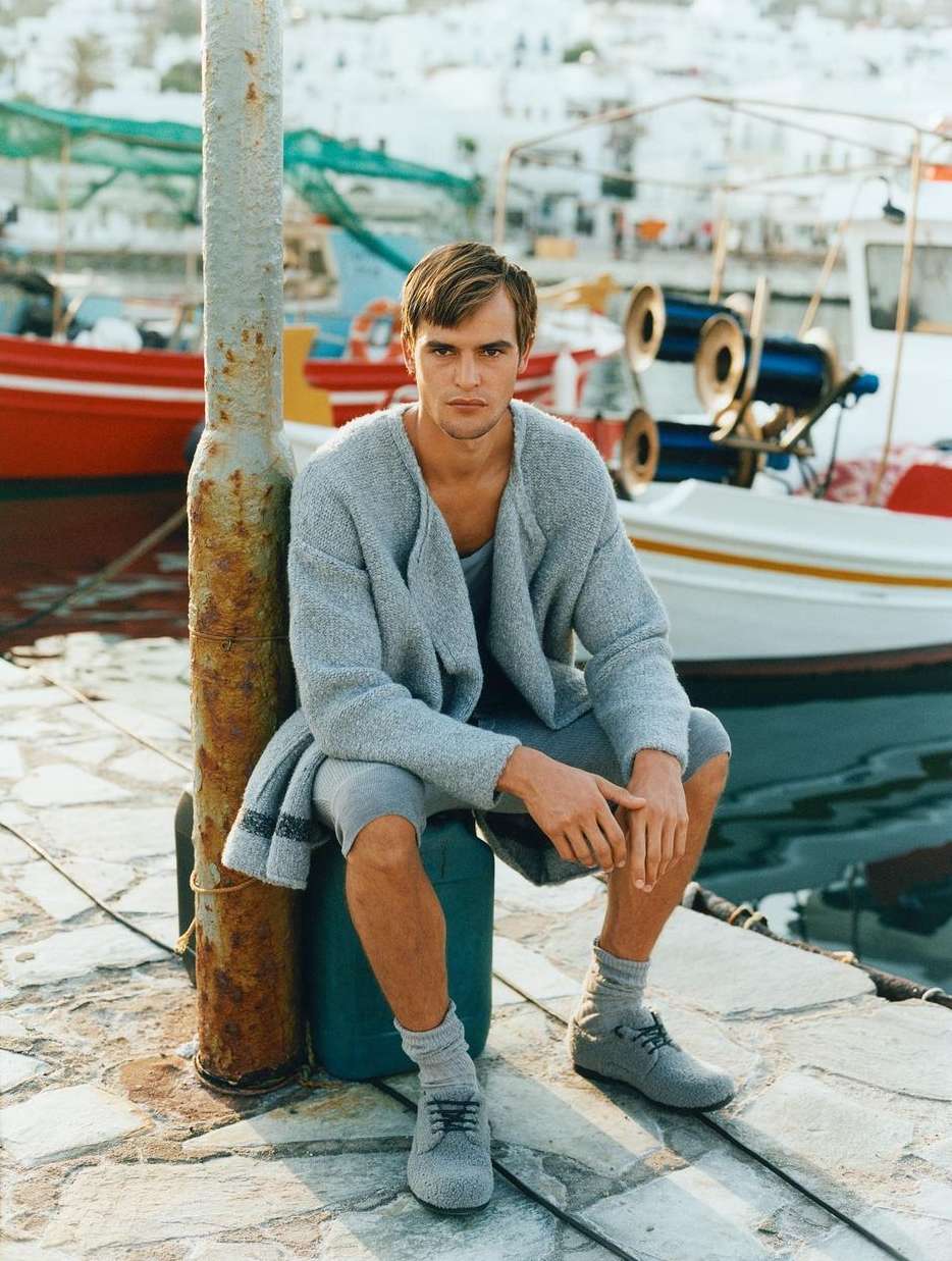 Young man in a grey knit outfit sits on a dock near fishing boats on a sunny day, with whitewashed buildings in the background.