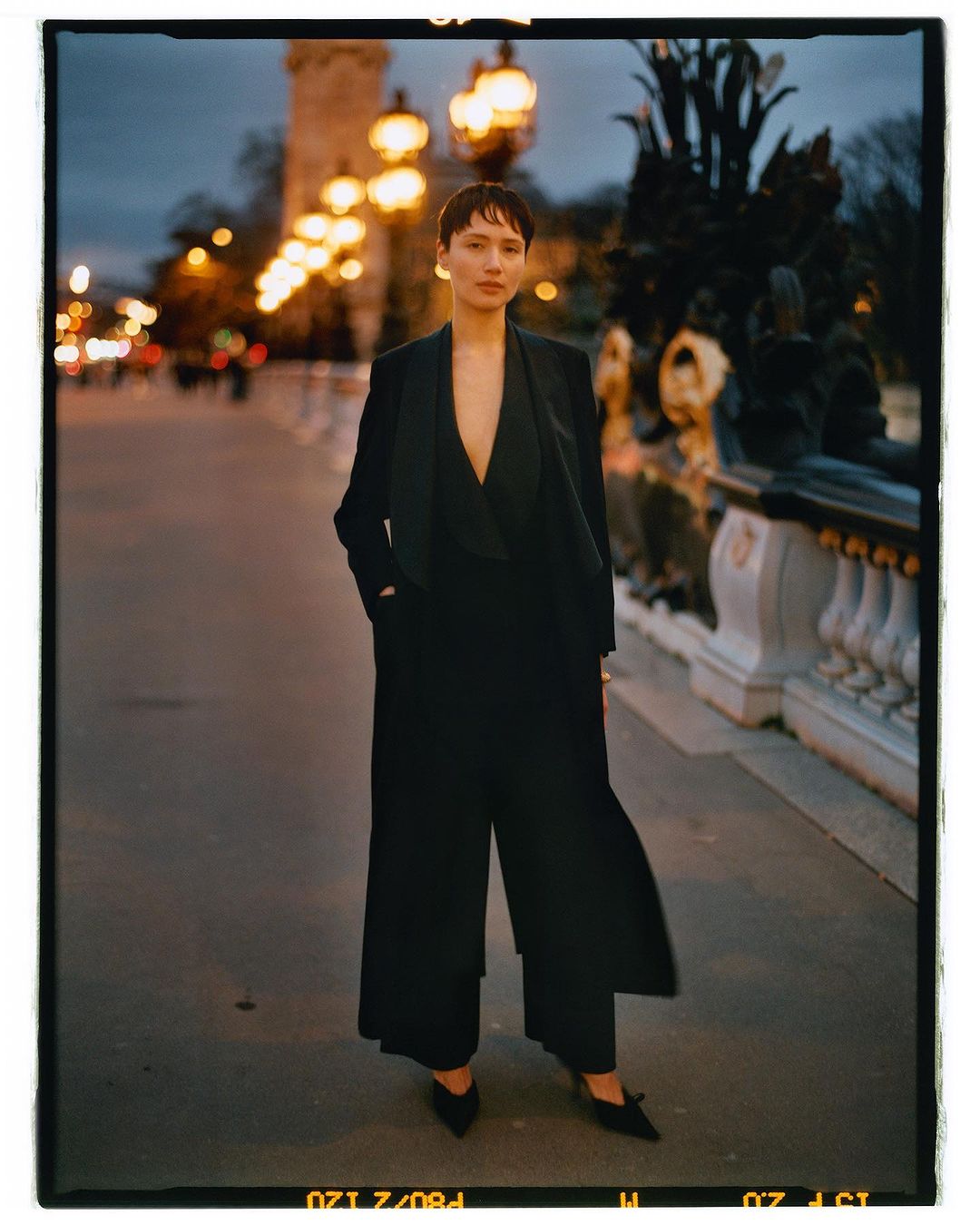 A woman in a black suit stands on a bridge with ornate railings and glowing streetlights in an outdoor nighttime fashion editorial.