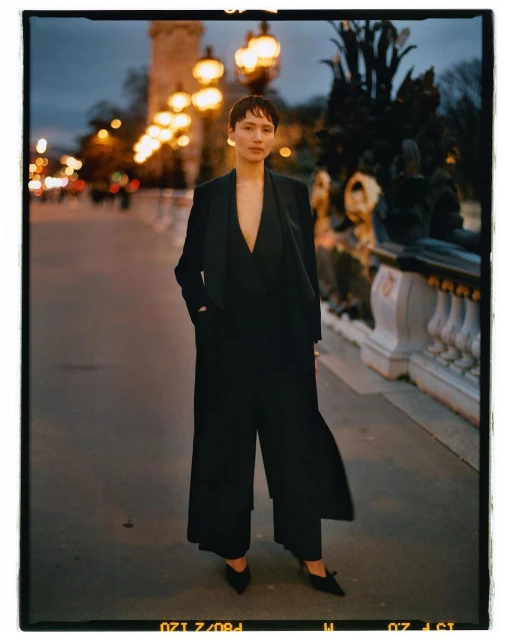 A woman in a black suit stands on a bridge with ornate railings and glowing streetlights in an outdoor nighttime fashion editorial.
