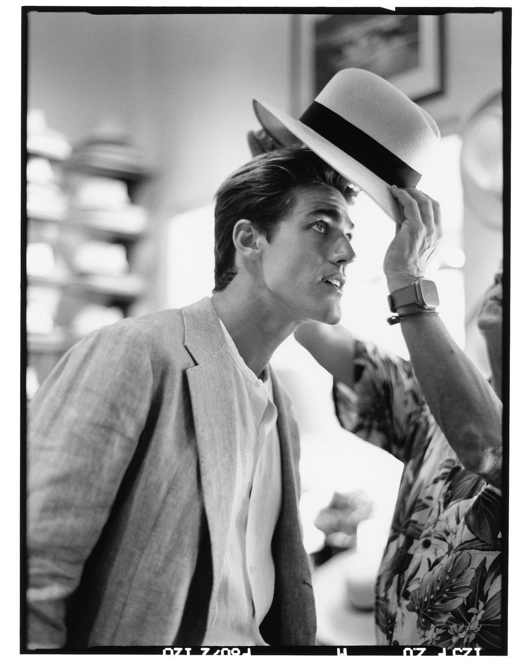 Monochrome shot of a stylish adult male getting fitted for a light-colored hat in a hat shop. The assistant wears a floral shirt.