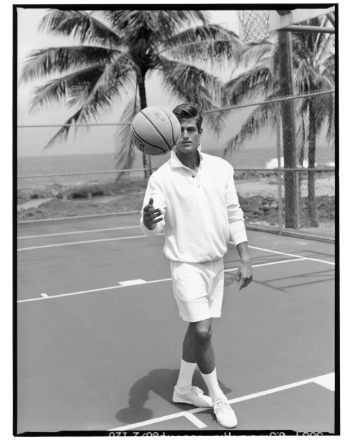 Black and white portrait of an adult male in athletic wear spinning a basketball on his finger, with palm trees in the background.
