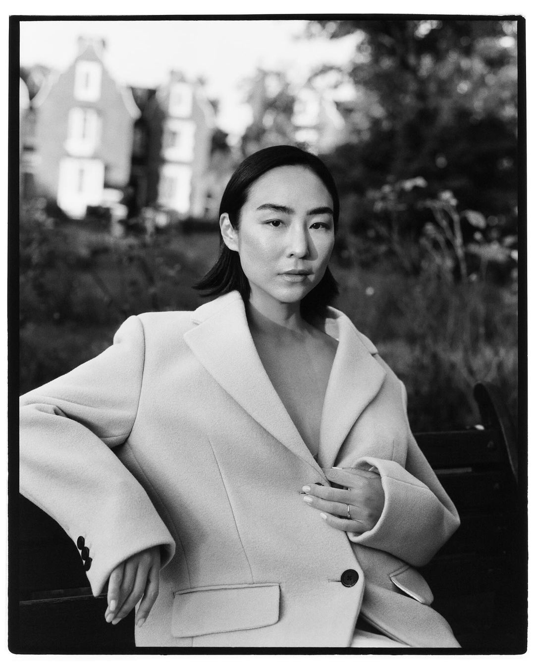Portrait of an Asian woman in a cream-colored blazer sitting on a bench outdoors, with soft focus on a building in the background.