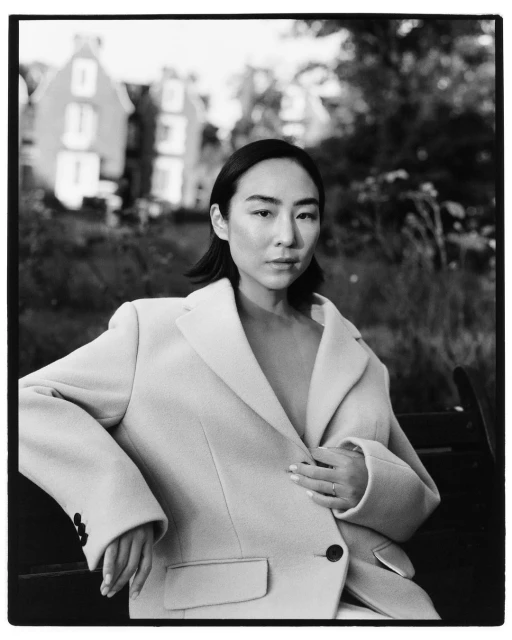 Portrait of an Asian woman in a cream-colored blazer sitting on a bench outdoors, with soft focus on a building in the background.