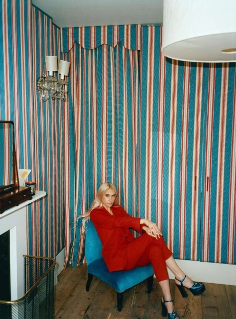 Woman in red suit poses on blue chair in striped interior. Stylish, fashion-forward portrait with vintage aesthetic.