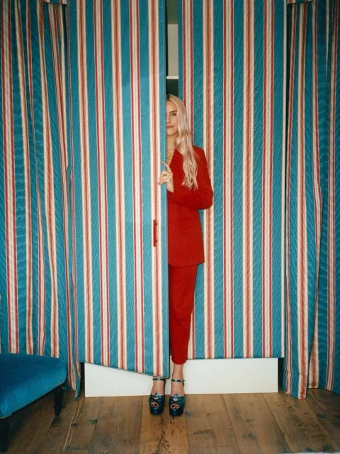 Woman with long blonde hair wearing a red suit peeking through striped blue, red, and white curtains in an indoor setting.