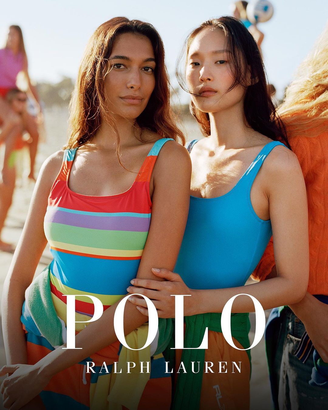 Two models pose on a sunny beach, wearing colorful swimsuits from Polo Ralph Lauren, with beachgoers visible in the background.