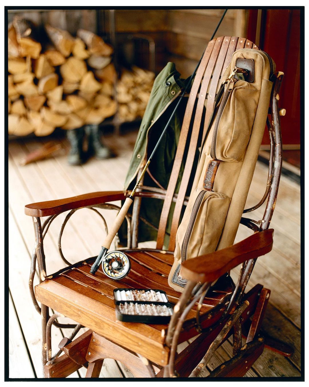 Fishing gear on a rustic wooden rocking chair: fly rod, reel, tackle box, vest. Woodpile and wooded outdoor setting.
