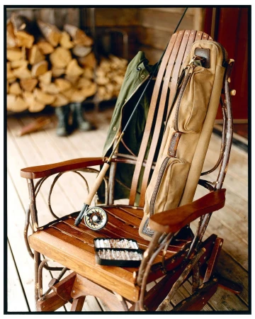 Fishing gear on a rustic wooden rocking chair: fly rod, reel, tackle box, vest. Woodpile and wooded outdoor setting.