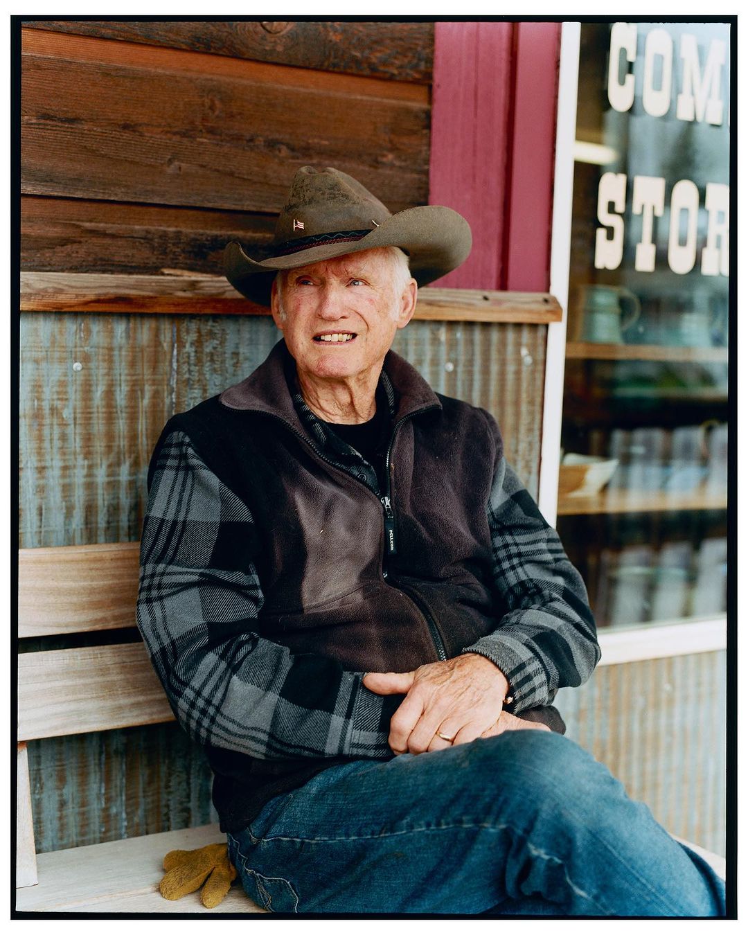 Elderly man in a cowboy hat sits on a wooden bench outside a shop with a weathered wood facade, wearing a plaid shirt and jeans.