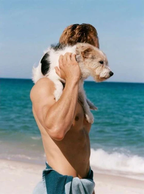 Athletic man carries a small dog on his shoulders at the beach with the turquoise ocean in the background on a sunny day.