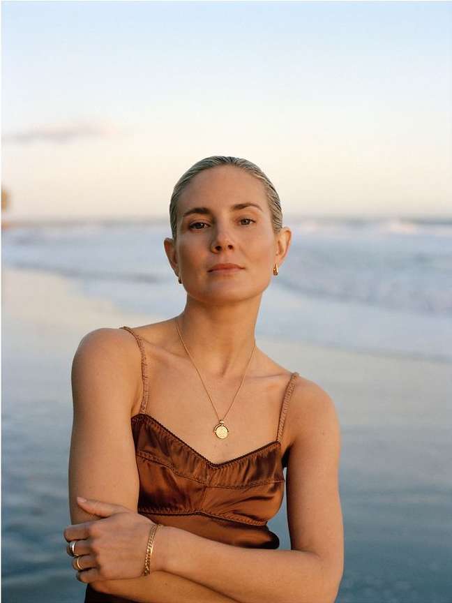 A woman with blonde hair poses on a beach in a brown dress with crossed arms, wearing jewelry and looking at the camera during daytime.