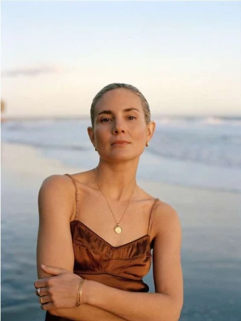 A woman with blonde hair poses on a beach in a brown dress with crossed arms, wearing jewelry and looking at the camera during daytime.