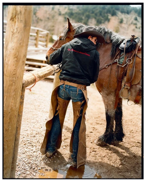 A horse and rider are shown in this outdoor shot. The rider is wearing chaps, a hat, and a jacket as they stand by the animal.