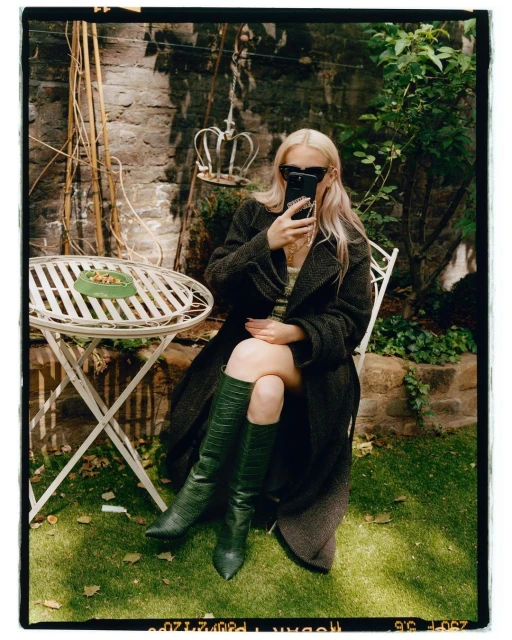 Blond woman in sunglasses takes smartphone photo while sitting in a coat and boots at a table outdoors on the grass.