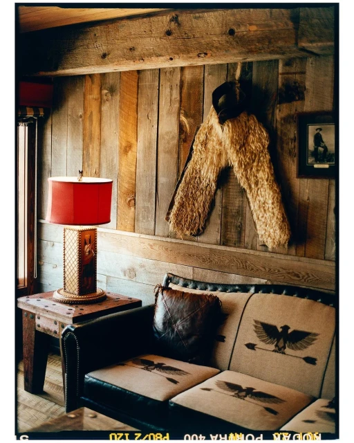 Interior of a rustic living room featuring a leather couch with eagle and arrow designs, a lamp, and a wooden wall with decorative chaps.