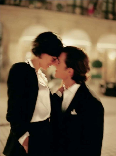 Romantic moment captured of a couple standing close with foreheads touching; the man is wearing a tuxedo and the woman a blazer.