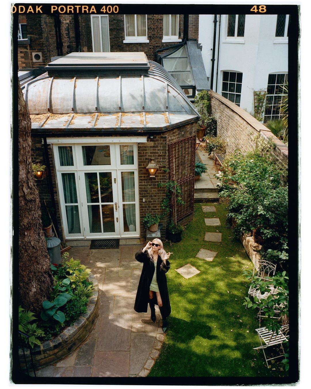Blonde woman in a long coat and sunglasses stands in a lush green backyard with stone pavers, foliage, and a sunroom. Shot from above.
