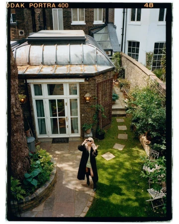 Blonde woman in a long coat and sunglasses stands in a lush green backyard with stone pavers, foliage, and a sunroom. Shot from above.