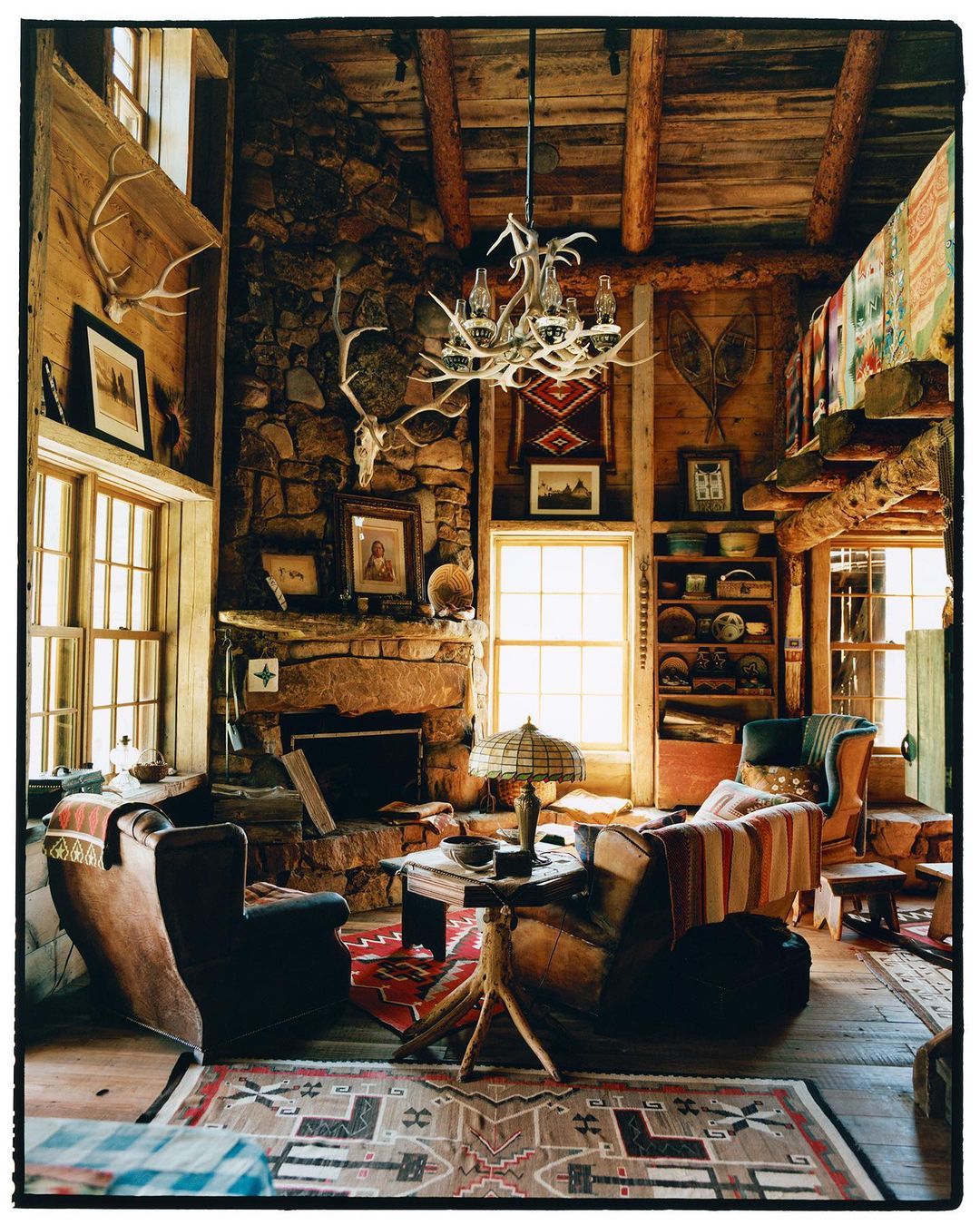 Cozy living room interior with stone fireplace, rustic wood walls, antler chandelier, and comfortable chairs under natural lighting.