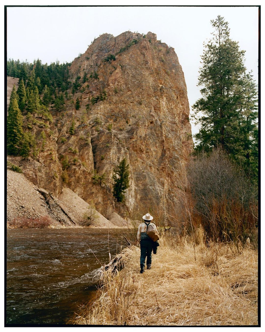 Man in hat walks along grassy path by river with large rock formation in the background on an overcast day outdoors.
