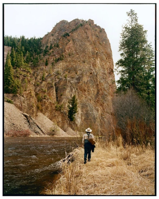 Man in hat walks along grassy path by river with large rock formation in the background on an overcast day outdoors.