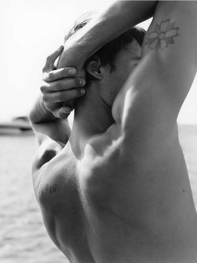 Monochrome portrait of a shirtless adult male with arm tattoos outdoors by the water, showcasing upper body muscles and shoulder blades.