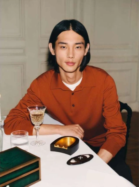 Portrait of a young Asian man seated at a table with drinks and accessories, styled in an indoor setting with a neutral background.