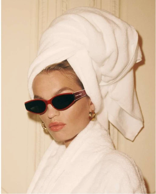 Adult woman wearing a white towel on her head and a white bathrobe with red sunglasses and gold hoop earrings in an indoor setting.