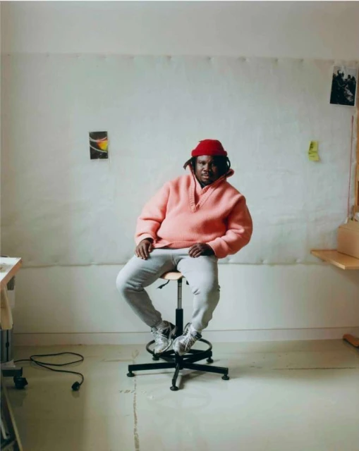 Indoor portrait of an African-American adult male sitting on a stool, wearing a pink hoodie, gray pants, white sneakers, and a red hat.