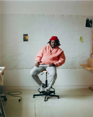 Indoor portrait of an African-American adult male sitting on a stool, wearing a pink hoodie, gray pants, white sneakers, and a red hat.