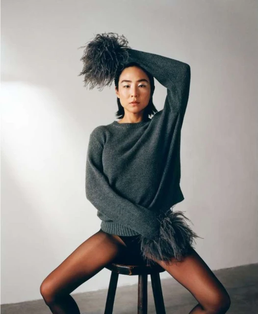 An Asian woman sits on a wooden stool in a gray sweater with feathered trim, styled with a feathered headband, against a white backdrop.
