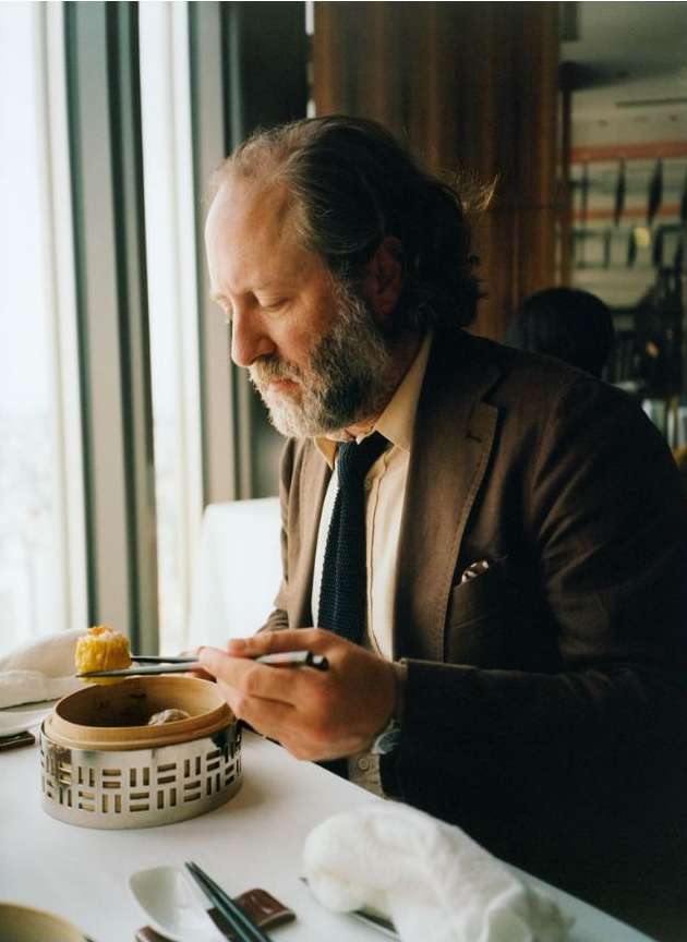 Adult male in brown suit jacket at a table with silver food warmer and chopsticks near a window with natural light in a restaurant.