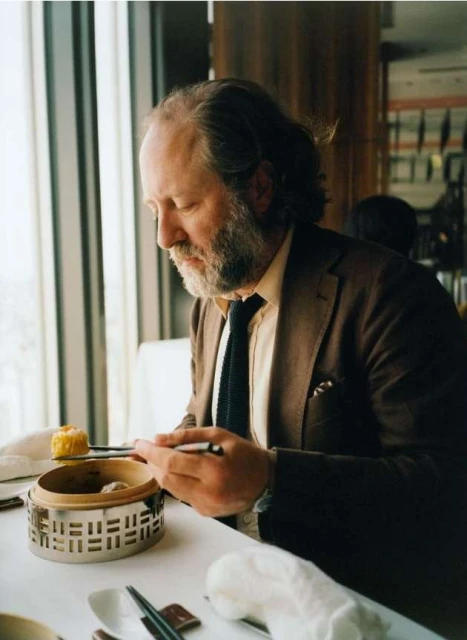 Adult male in brown suit jacket at a table with silver food warmer and chopsticks near a window with natural light in a restaurant.