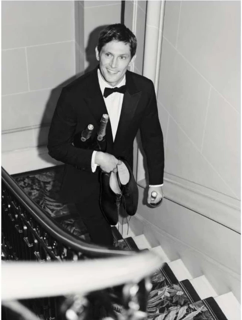 Black-and-white photo of a man in a tuxedo carrying wine and shoes on a staircase with a smile on his face. Formal attire.