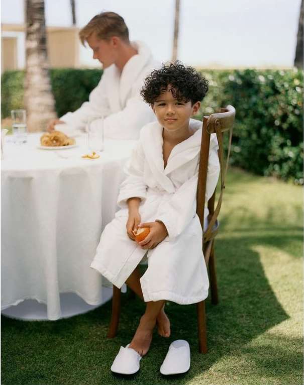Child and young adult wearing white robes, seated at a table on a lawn with a building and palm trees in the background on a sunny day.