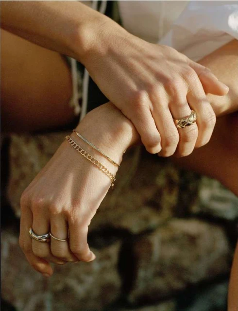 Close-up of a woman's arm and hand displaying fashionable rings and gold bracelets, set against an out-of-focus backdrop.