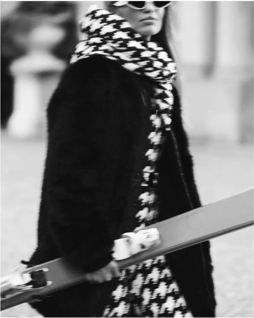 Stylish woman in a black coat, patterned scarf, sunglasses, and skis walking outdoors in a black and white fashion photograph.